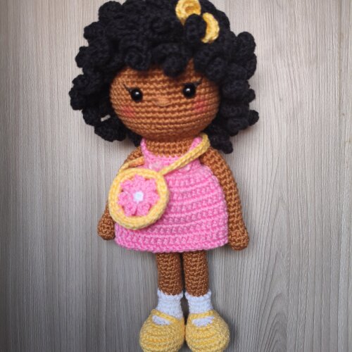 Boneca amigurumi com vestido cor de rosa, bolsinha e lacinho amarelo com cabelos encaracolados
